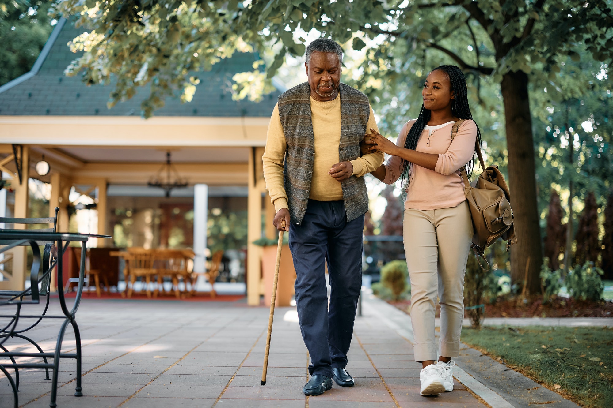 Black senior man with walking cane and his daughter a taking walk through park of nursing home.